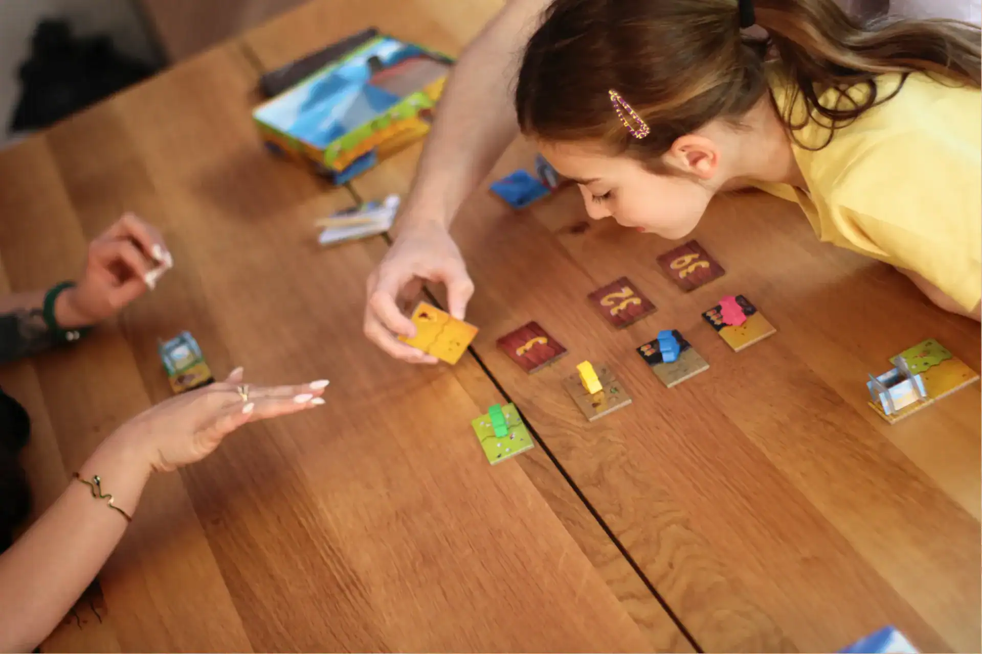 Zwei Personen spielen ein Brettspiel mit Karten, Figuren und Spielmaterial auf einem Holztisch.